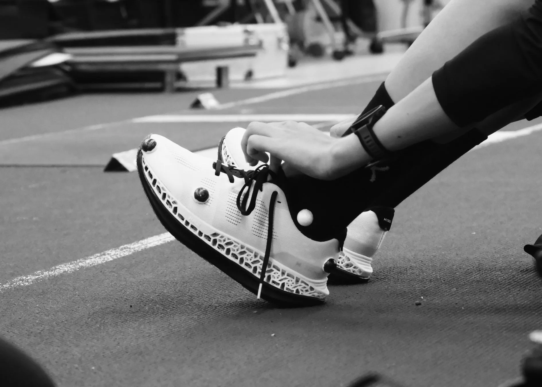 アスリートが靴紐を結んでいるモノクロ写真。アスファルトの上で、白いスニーカーを履いた足が写っており、手で紐を結んでいる様子が捉えられている。背景には他の人や器具の一部がぼやけて写っている。