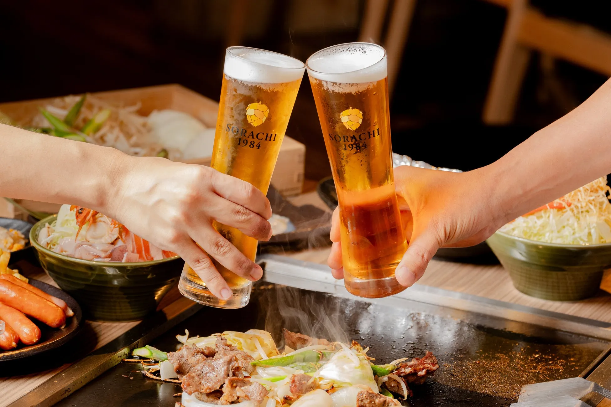 ビールグラスを乾杯する様子、鉄板焼きの料理と様々な食材が写っている。