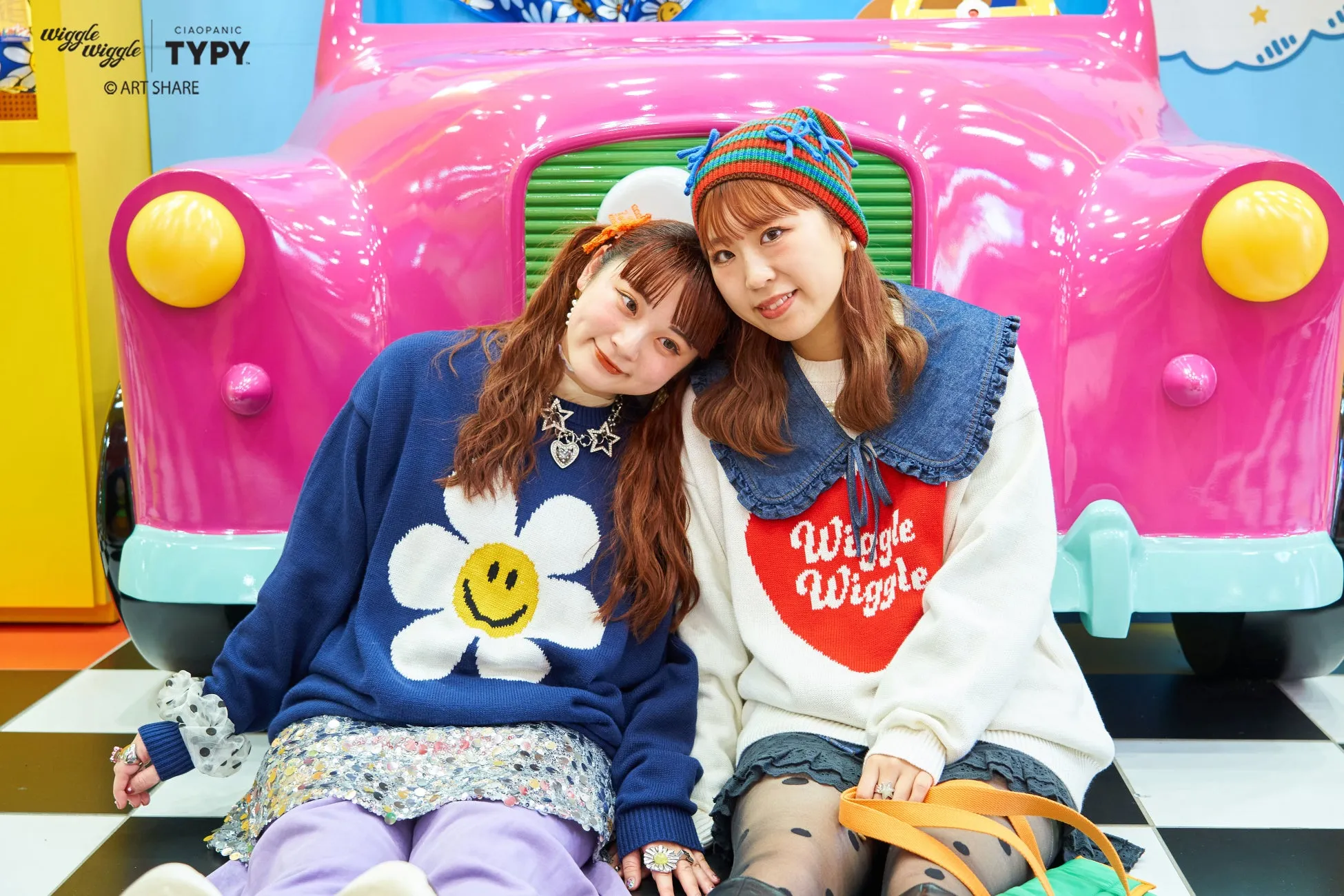 ピンクの車のオブジェの前で寄り添う2人の女性。左の女性はスマイルマークのセーター、右の女性はハートに「Wiggle Wiggle」と書かれたセーターを着ています。