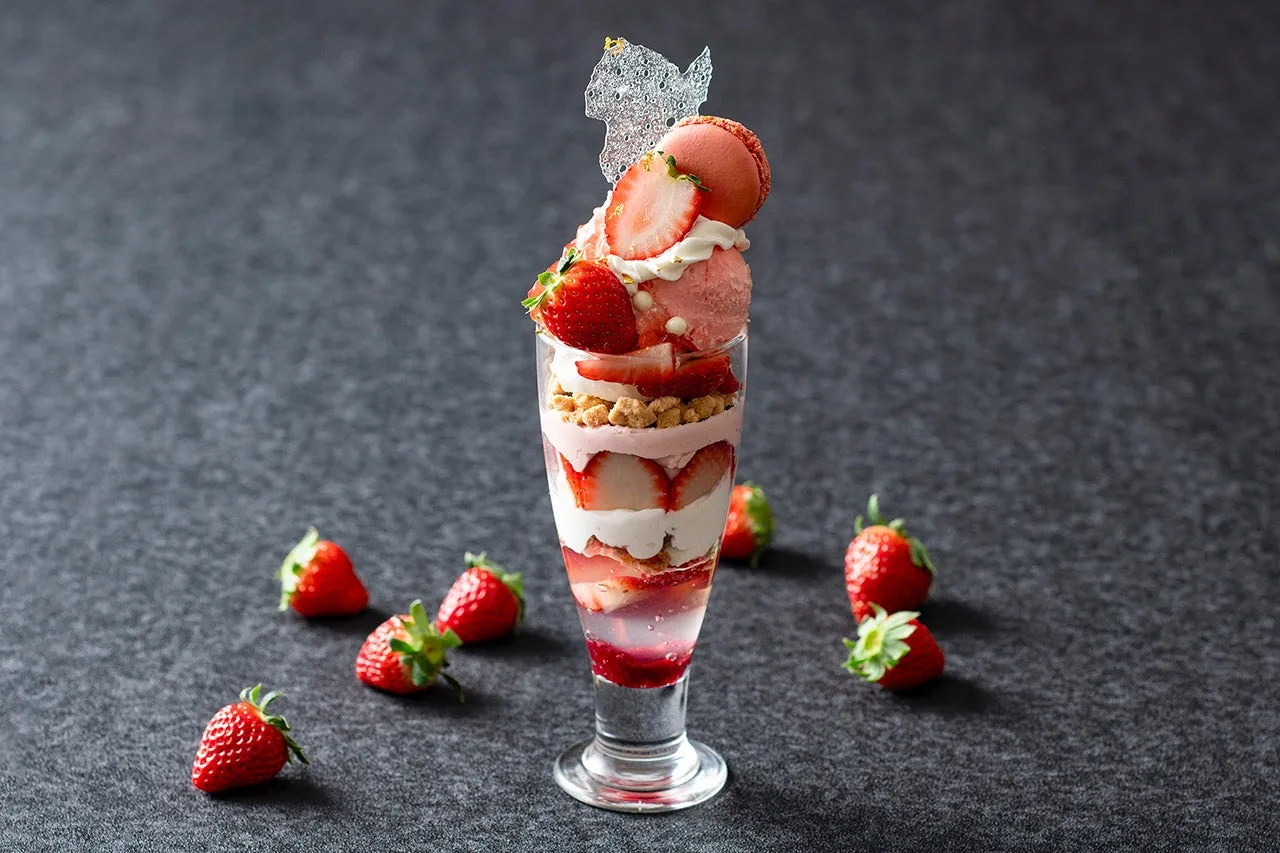 グラスに入ったイチゴパフェの写真。パフェは、イチゴ、クリーム、クランブル、アイスクリーム、マカロンなどで構成されている。周りにはイチゴが散りばめられている。