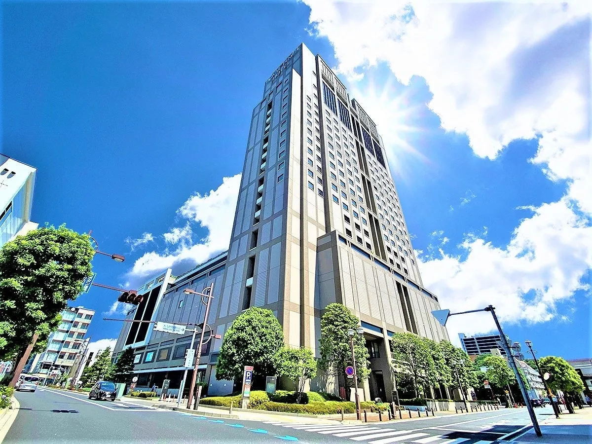 青空の下、高層ビルがそびえ立つ様子を捉えた写真。建物の下部には緑豊かな木々が植えられ、道路には横断歩道や信号機が見えます。