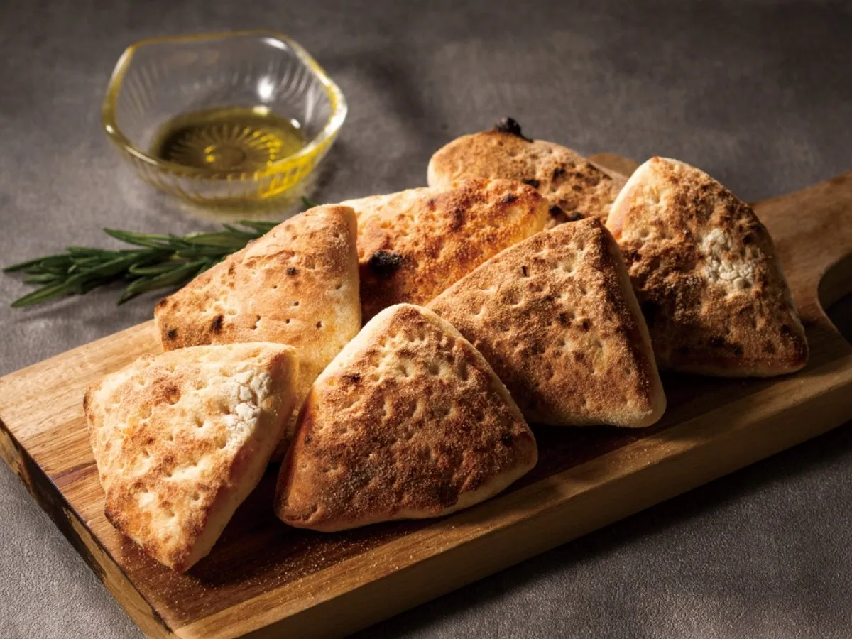 木製のまな板に三角形のパンが並べられ、オリーブオイルとローズマリーが添えられています。