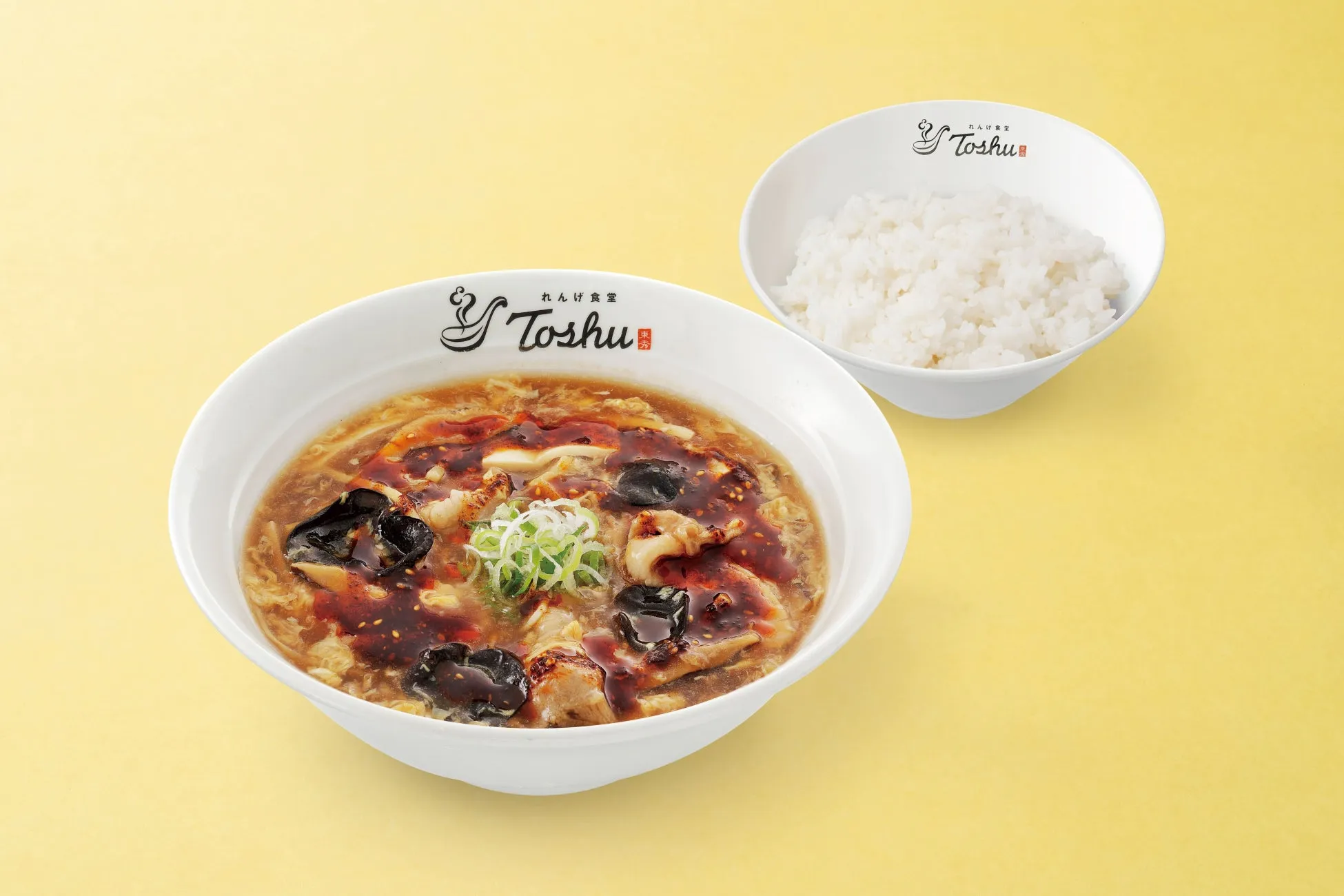 白い器に入ったラーメンとご飯の写真。ラーメンは、具材が豊富で、スープが赤茶色。ご飯は白く、シンプルな盛り付け。背景は黄色。