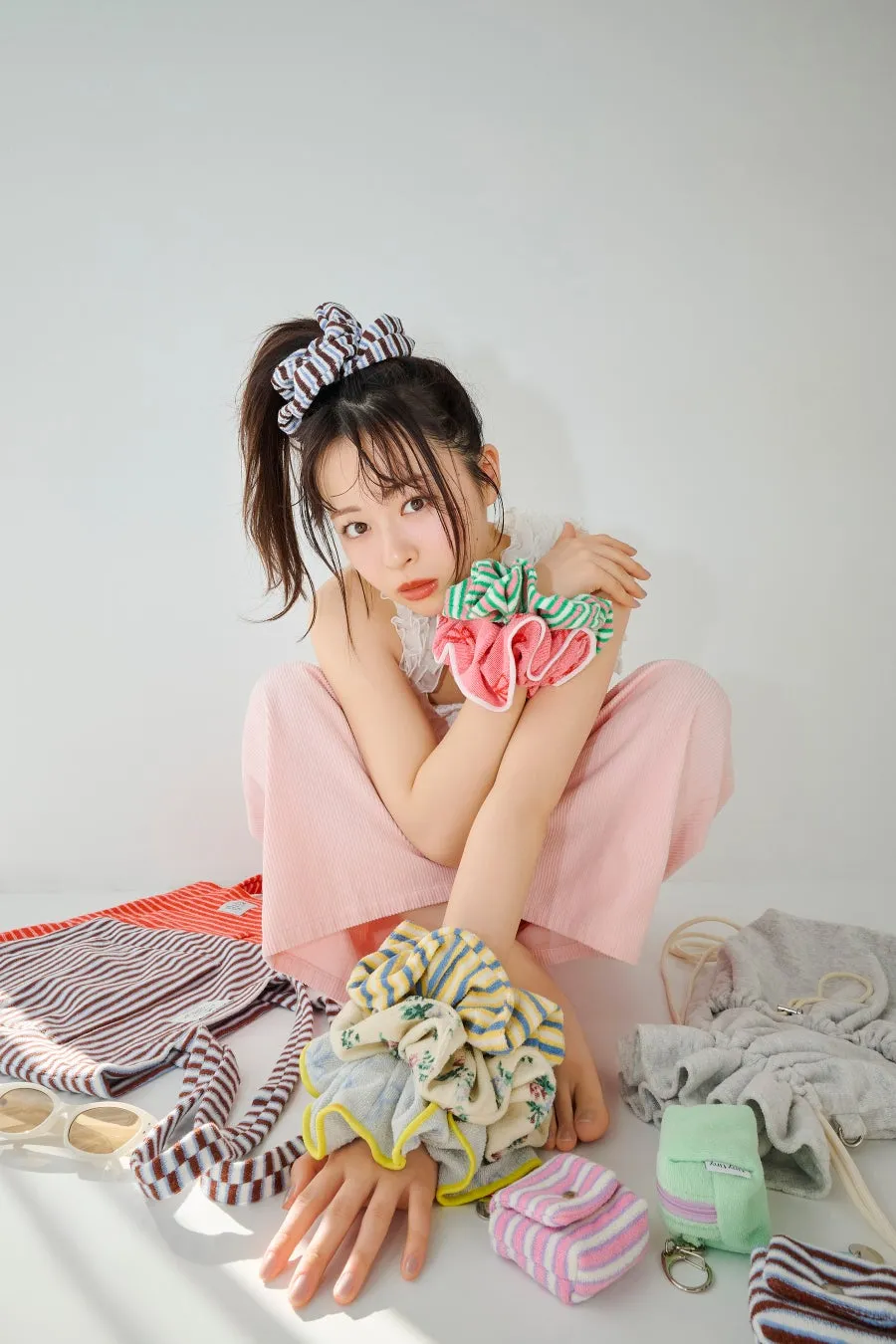 女性が様々な柄のシュシュやバッグ、サングラスなどの小物を身につけ、床に座ってポーズをとっている様子。