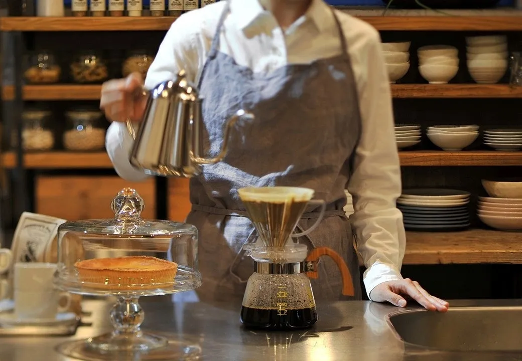 コーヒーを淹れている様子。店員がケトルからドリッパーに湯を注いでいる。手前にはケーキが置かれ、棚には食器が並んでいる。