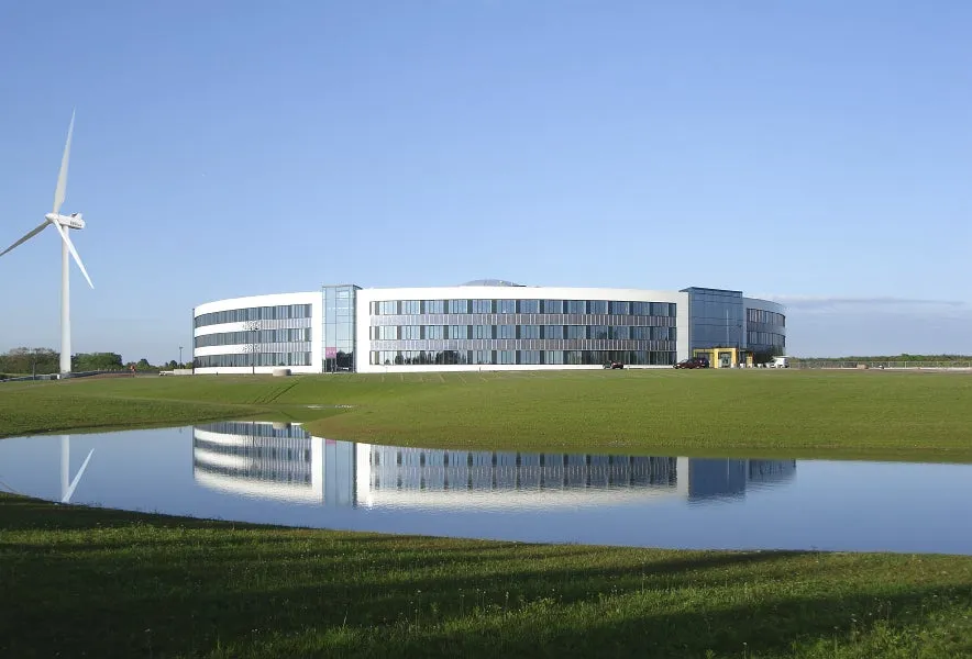 風力タービンと、水面に反射するモダンなオフィスビルがある風景写真です。