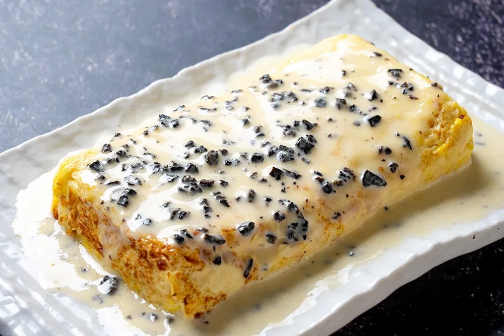 白い長方形の皿に盛られたオムレツ。オムレツは黄色く、白いソースで覆われ、黒いトッピングが散りばめられている。