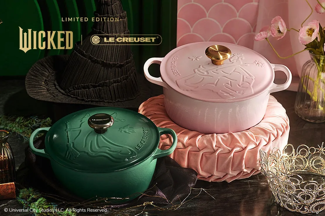 2つの鍋と帽子、王冠、花瓶が置かれた写真。鍋は緑とピンクで、それぞれに魔女の絵が描かれています。背景には、映画「Wicked」のロゴと、Le Creusetのロゴがあります。