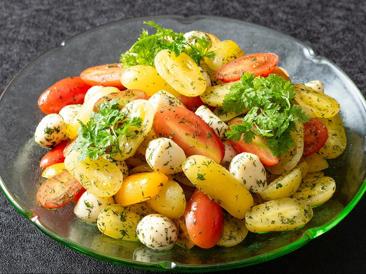 透明なガラスの皿に盛られた、黄色と赤のトマト、白いチーズ、パセリのサラダ。