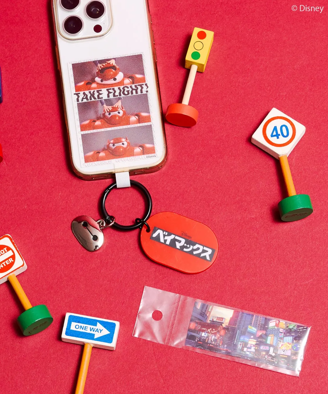 赤い背景に、ベイマックスのキャラクターが描かれたスマホケース、キーホルダー、信号機や道路標識の玩具が配置された写真