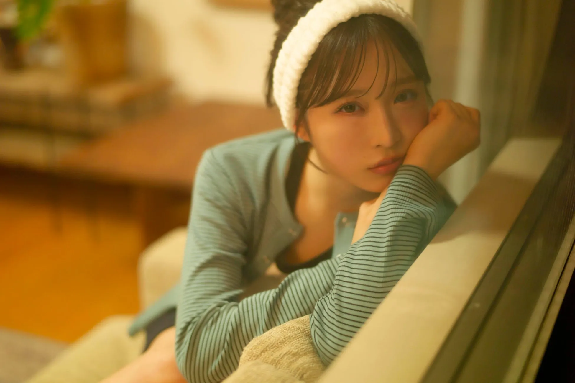 女性がソファに寄りかかっていて、手で頬杖をついている。白いヘアバンドとストライプのシャツを着ている。