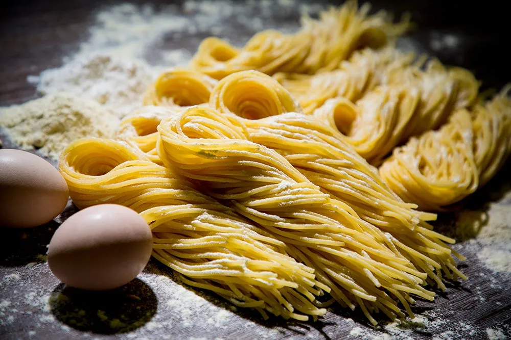 パスタのクローズアップ写真。パスタは、卵と小麦粉の周りに円形に積み重ねられています。表面には小麦粉がまぶされています。