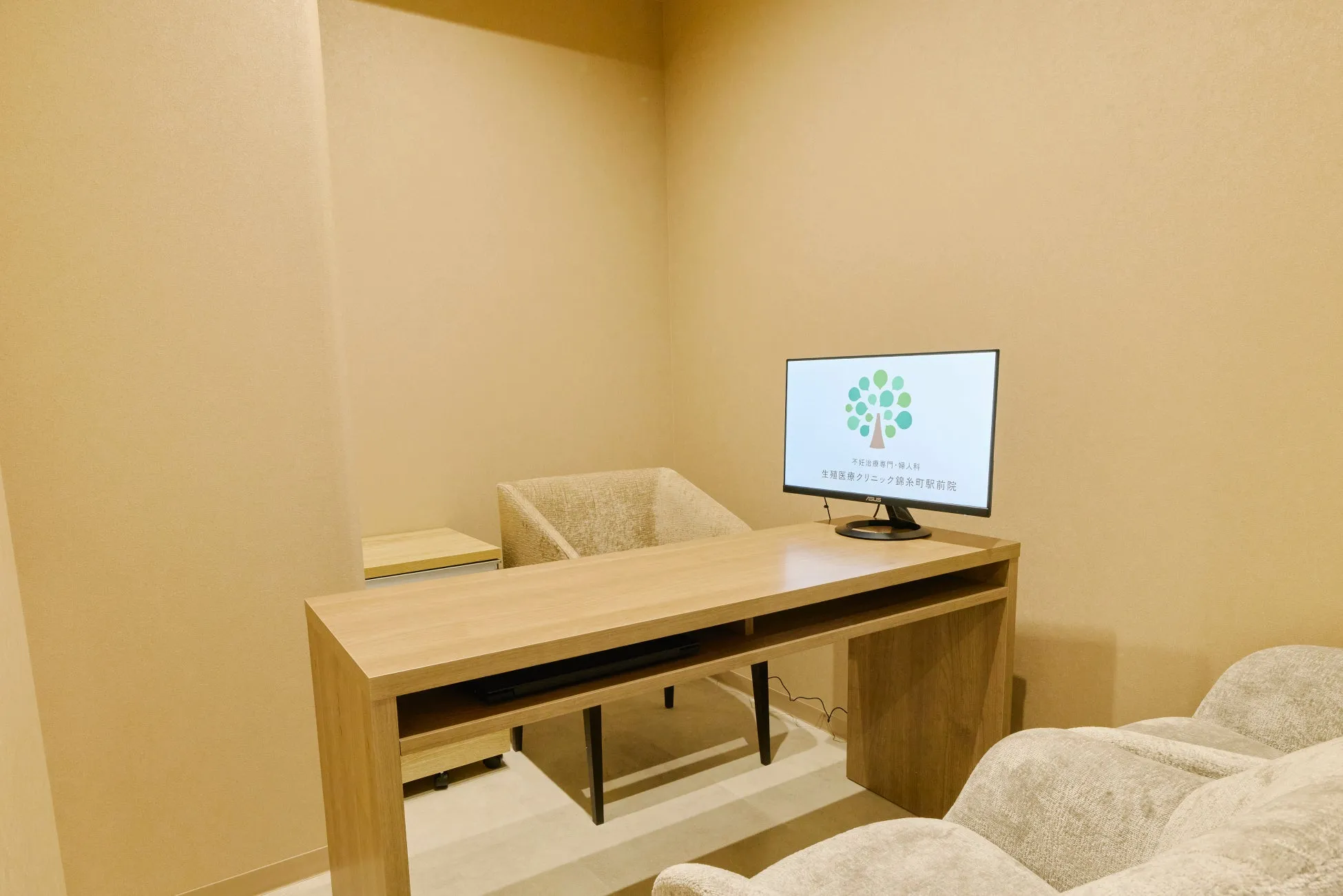 クリニックの診察室の写真です。木目調のデスクとモニターがあり、壁はベージュ色です。