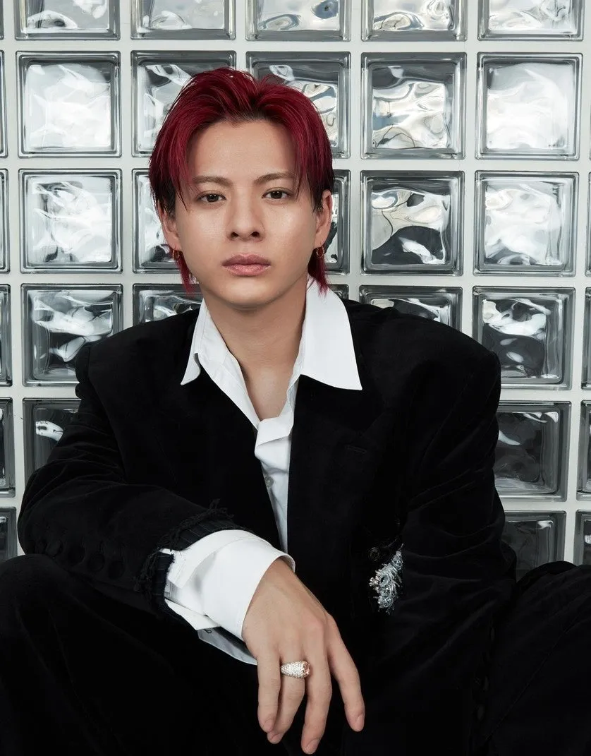 赤い髪の男性がカメラ目線で座っている姿。黒いスーツと白いシャツを着用し、指には指輪をしている。