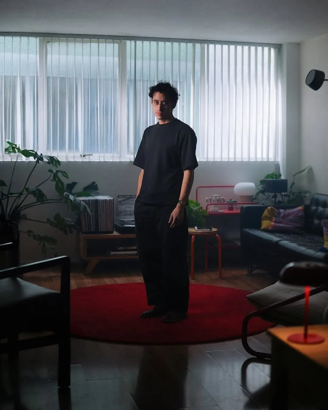 部屋に立つ男性の写真。彼は黒い服を着ており、カメラ目線で立っている。部屋にはレコードプレーヤー、ソファ、赤いカーペット、窓がある。