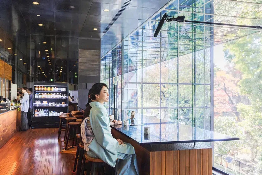 カフェで着物を着た女性が窓の外を眺めている様子。窓の外には紅葉した木々が見え、店内にはマイクや飲み物などが写っています。