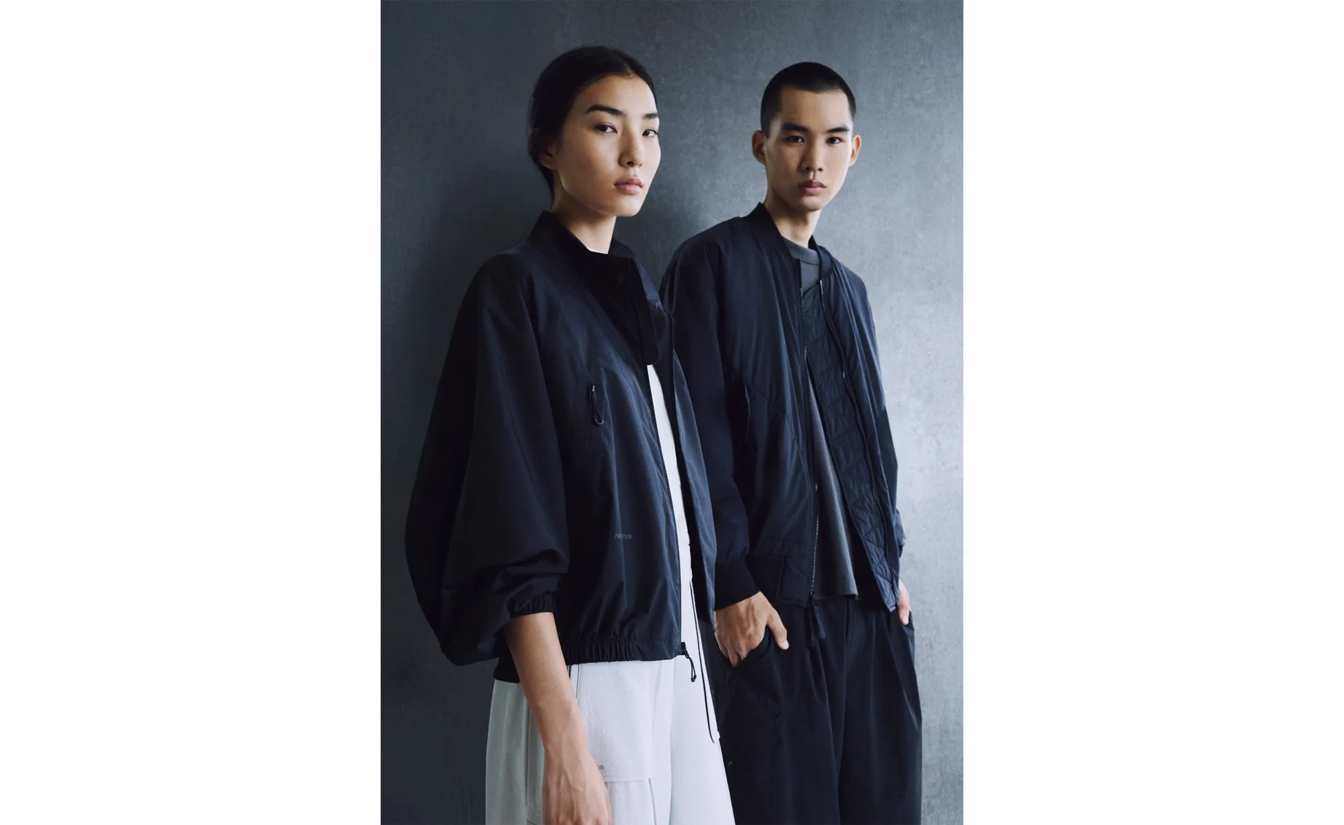 2人のモデルが黒いジャケットを着てポーズをとっている写真。女性は白いパンツ、男性は黒いパンツを履いています。背景はグレーの壁です。