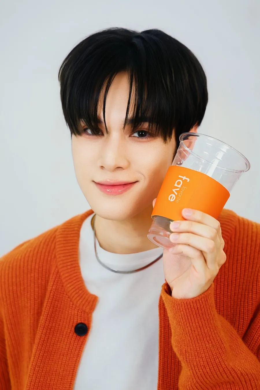 オレンジ色のカーディガンを着た男性が、faveのロゴが入ったカップを持っています。彼はカメラ目線で微笑んでいます。