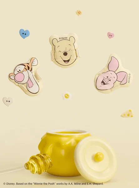 くまのプーさんのキャラクター、ティガー、プーさん、ピグレットの顔の形をしたクッション、はちみつポット、はちみつスプーンが配置された画像。