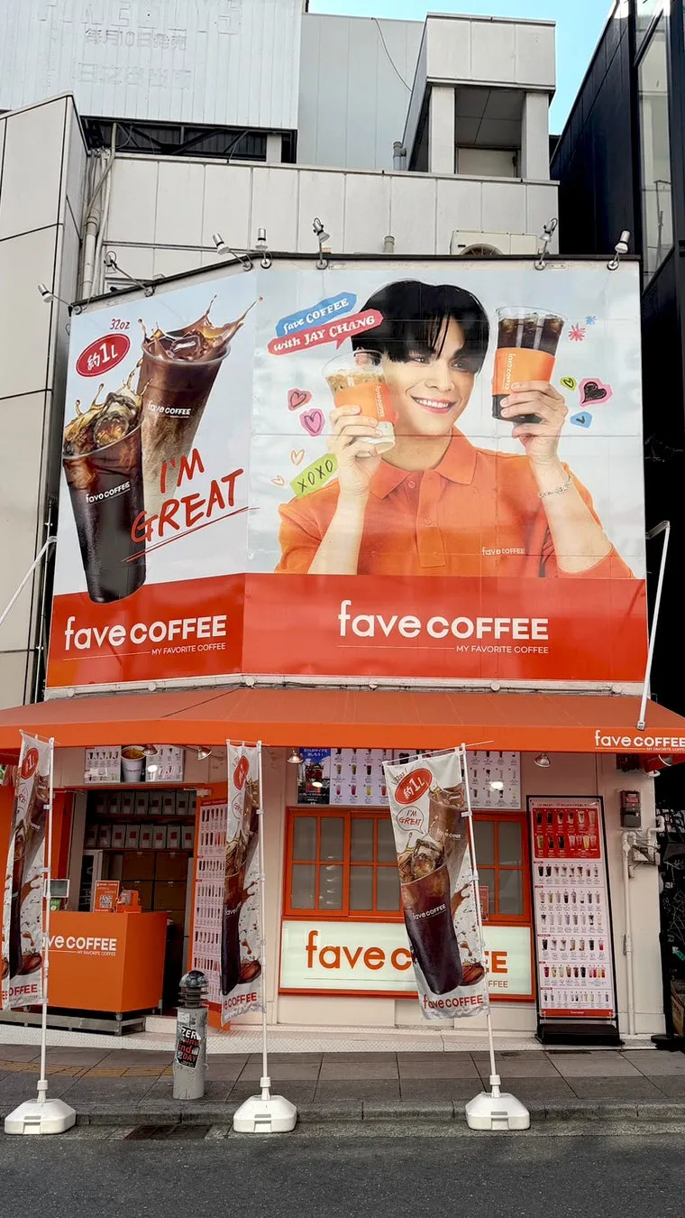 カフェ「fave COFFEE」の屋外広告と店舗の写真。広告には、JAY CHANGがコーヒーカップを持ち笑顔で写っており、「Save COFFEE with JAY CHANG」という文字とコーヒーの写真が描かれている。店舗はオレンジ色を基調とし、コーヒーのメニューやロゴが掲示されている。