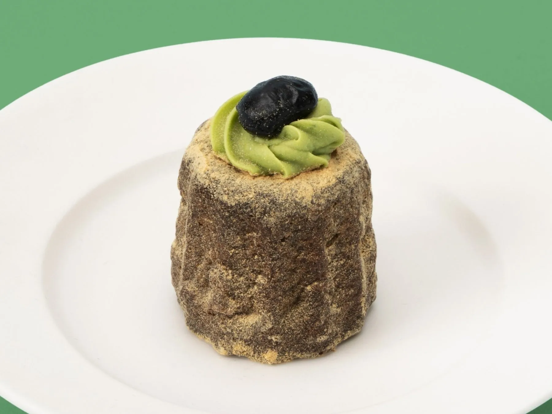 白い皿の上に、抹茶クリームと黒い実がトッピングされた小さな焼き菓子が置かれています。背景は緑色です。