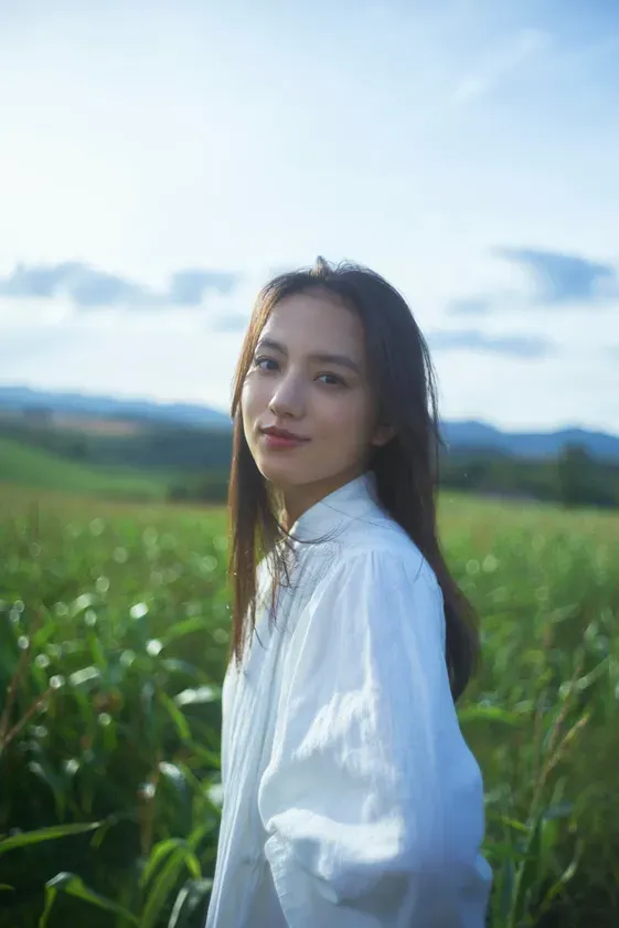 白いシャツを着た女性が、緑の畑を背景に微笑んでいます。空は青く、太陽の光が差しています。