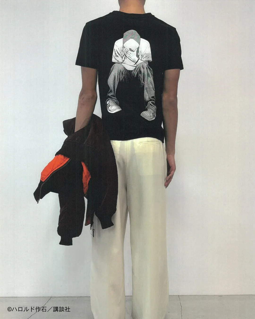 黒いTシャツに座っている男性のイラストが描かれており、白いパンツを履いている。男性は、片手にジャケットを持っている。
