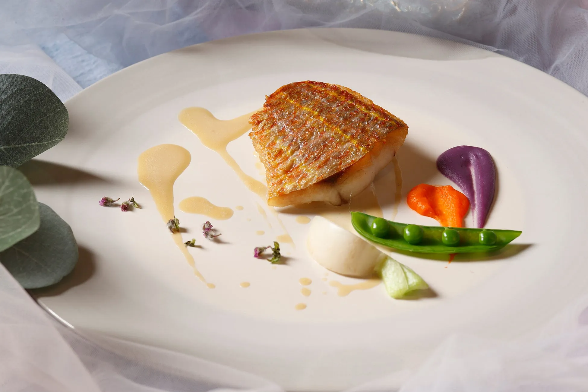 白い皿に盛られた、焼き魚、ソース、野菜の料理。彩り豊かで、芸術的な盛り付けが特徴的。