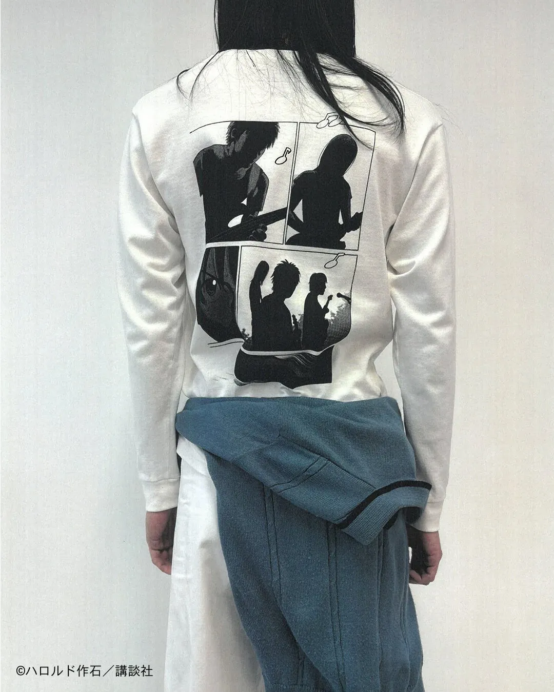 長袖Tシャツを着た人物の後ろ姿。Tシャツには、漫画のコマ割りのように、ギターを演奏する人物のシルエットが描かれている。人物は白い壁を背景に立っている。
