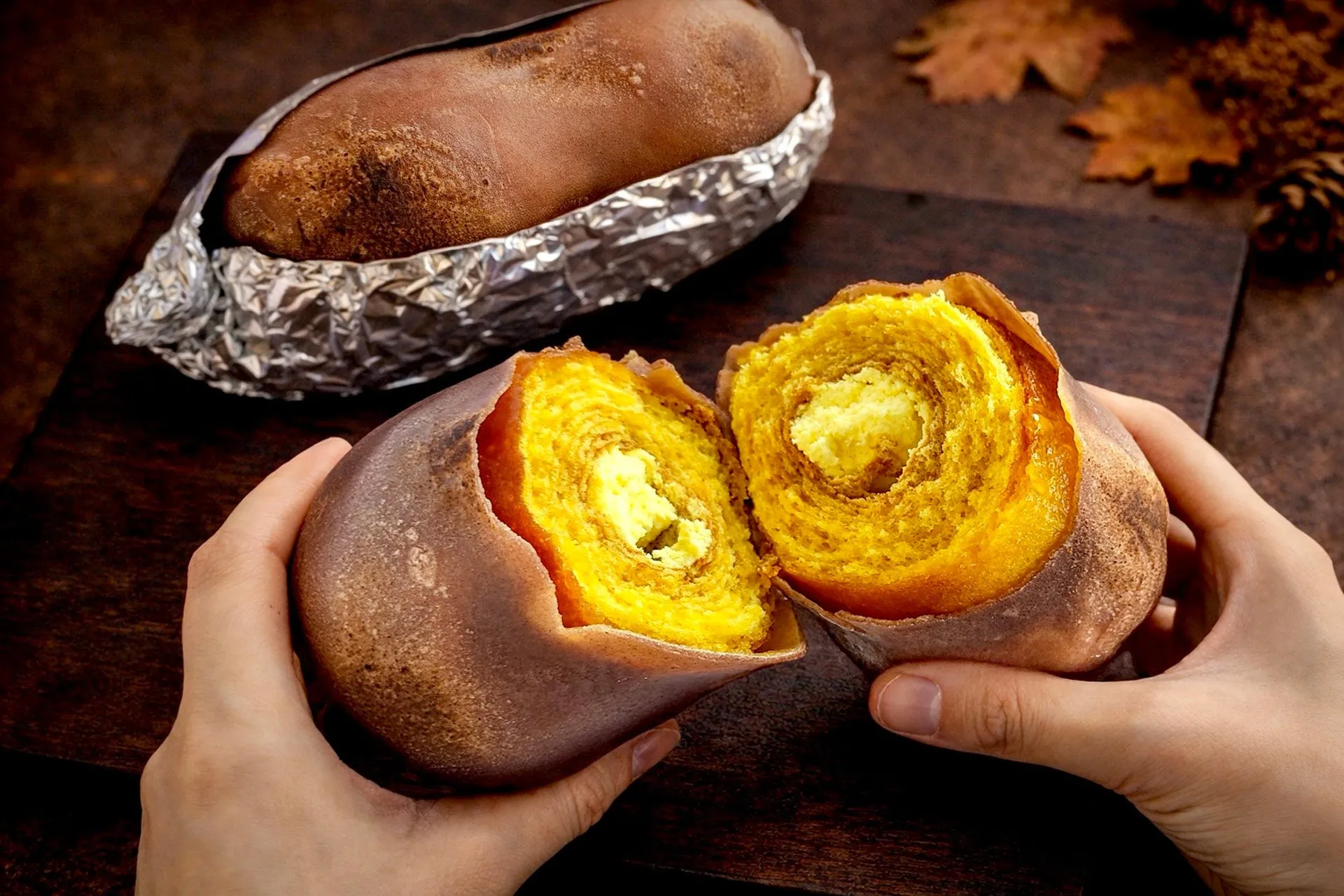焼き芋を切った断面と、それを持つ手元を写した写真。焼き芋はアルミホイルに包まれており、断面は黄色く、年輪のような模様が見える。背景には木製の板や紅葉が写っている。