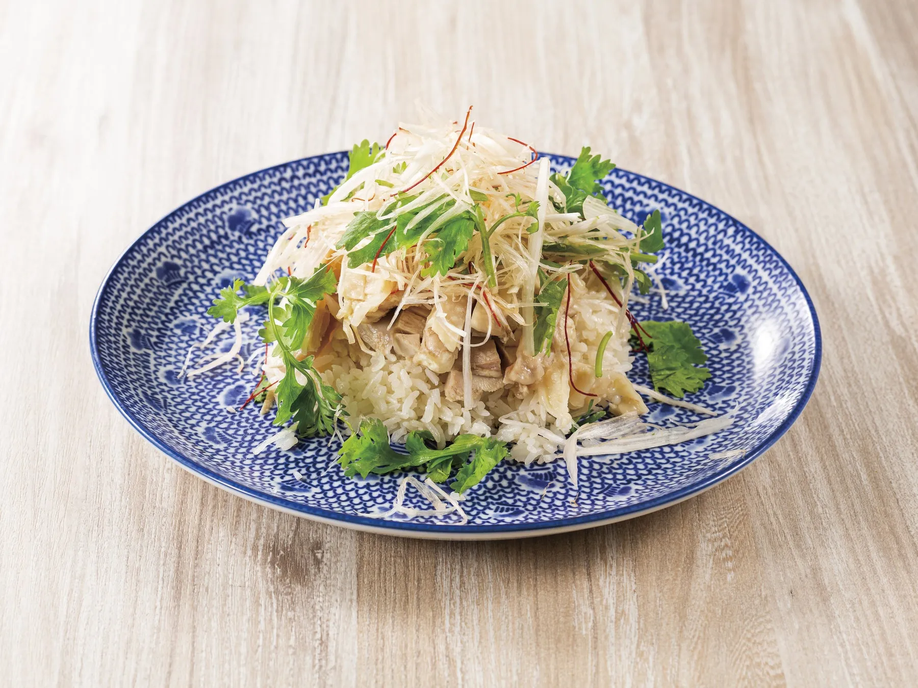 白いご飯の上に鶏肉と薬味が乗った料理が、青い模様の皿に盛られています。皿は木目のテーブルに置かれています。