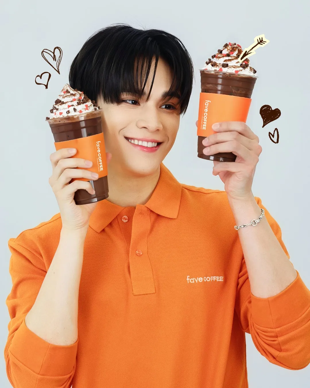 オレンジ色のポロシャツを着た男性が、2つのチョコレートドリンクを持ち、笑顔でポーズをとっている画像。ドリンクにはホイップクリームとトッピングが施されており、手書きのハートマークが描かれている。