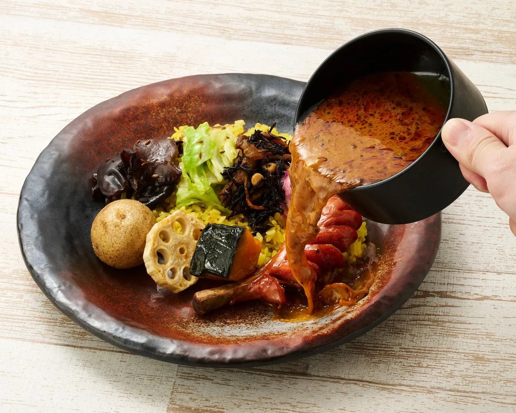 カレーがかけられる直前の料理の写真です。皿には、ターメリックライス、野菜、ソーセージが盛り付けられており、カレーが注がれている様子が写っています。