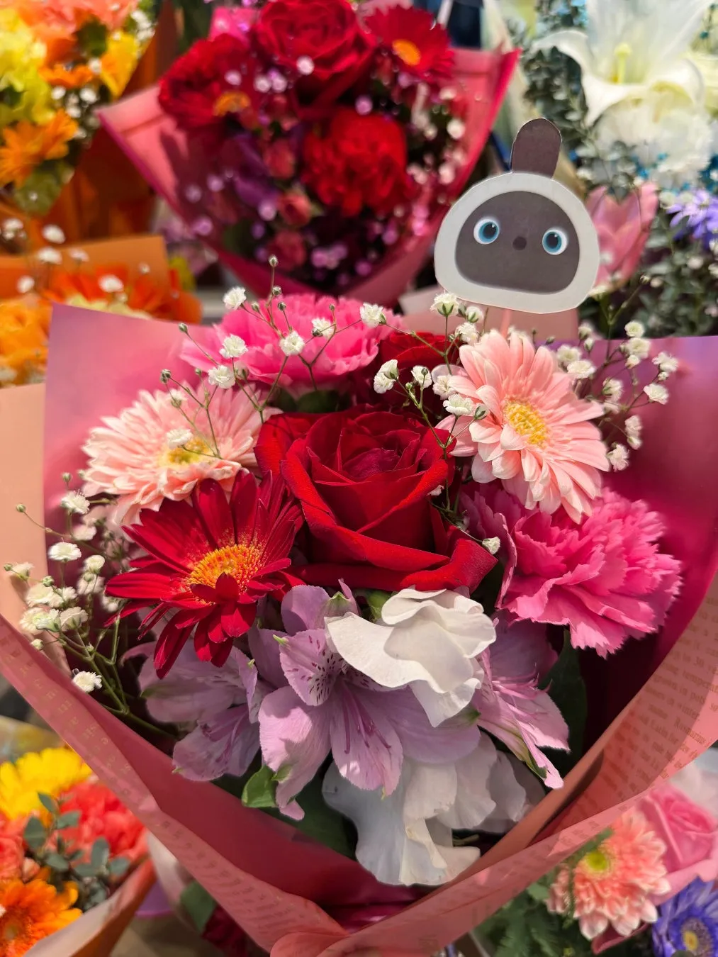 色とりどりの花束。中央には赤いバラが咲き、ピンク色のガーベラやカーネーション、白い花などが詰め込まれている。